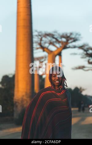 Madagascar - 6 LUGLIO 2019: Contenuto donna africana in esotica copertura multicolore con cappuccio in piedi da alberi di baobab in raggi solari al crepuscolo Foto Stock