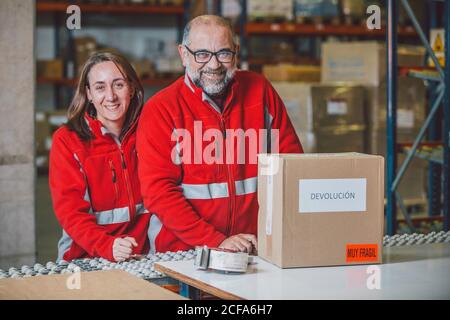 Soddisfatti dipendenti di magazzino maschi e femmine in uniformi rosse sorridendo e guardando la fotocamera con il pacchetto sul tavolo Foto Stock