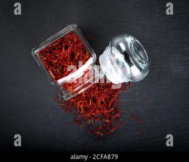 Vista dall'alto dello zafferano aromatico naturale essiccato versato dal vetro vasetto posizionato su un tavolo nero Foto Stock