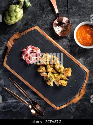 Composizione vista dall'alto con prosciutto a fette e carciofi cotti serviti su tavola di ardesia nera sul tavolo con salsa e posate Foto Stock