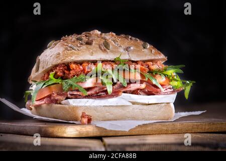 Primo piano di delizioso sandwich fatto con pane integrale ripieno di pancetta croccante e formaggio con pomodori freschi e lattuga serviti su tavola di legno su sfondo nero Foto Stock