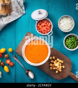 Vista dall'alto della ciotola del gazpacho andaluso con gustosi cracker in composizione con verdure fresche tritate e cipolla come ingredienti su tavolo di legno blu chiaro Foto Stock