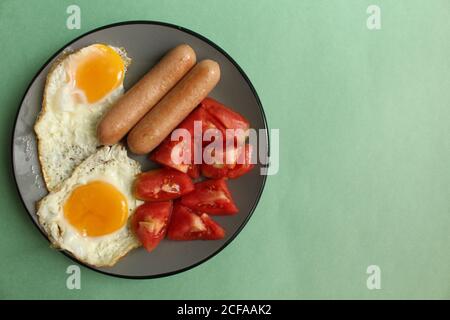 La colazione viene servita su un piatto grigio su sfondo verde salsicce pomodori uova strapazzate uova fritte con sfondo copyspace Foto Stock