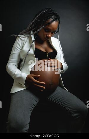 Giovane donna afro-americana incinta con trecce con blazer bianco e i jeans stroking pancia mentre si siede in studio contro nero parete Foto Stock