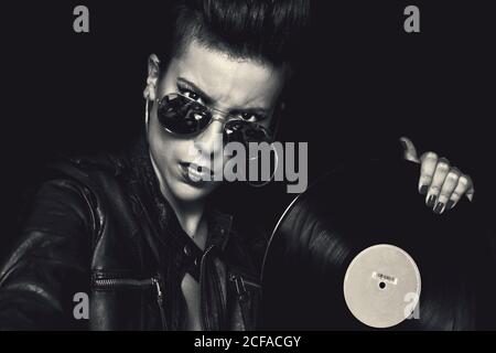 Arrabbiato sicuro femmina rocker in giacca di pelle e occhiali da sole aviatore guardando la macchina fotografica e tenendo il disco in vinile in studio sopra sfondo nero Foto Stock