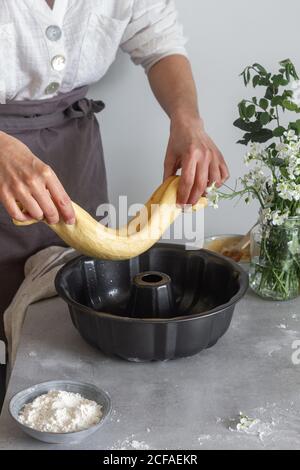 donna irriconoscibile nel grembiule che mette il rotolo di pasta morbida in Tegola vicino alla farina e ai fiori mentre prepari la torta Bundt tabella Foto Stock