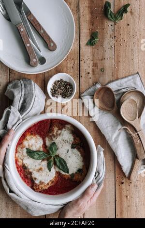 Vista dall'alto di una persona anonima che mette la ciotola calda piena di Delizioso pollo con parmigiano e foglie di basilico tostate in pomodoro salsa sul tavolo di legname vicino a spezie e utensili Foto Stock