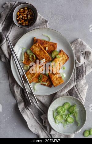 Vista dall'alto del delizioso pasto vegetariano, tofu fritto che marina salsa di soia con aglio e servire in piatto bianco spruzzando con pezzi di cipolla verde in piedi su asciugamano da cucina Tavolo grigio con bastoncini cinesi in acciaio Foto Stock