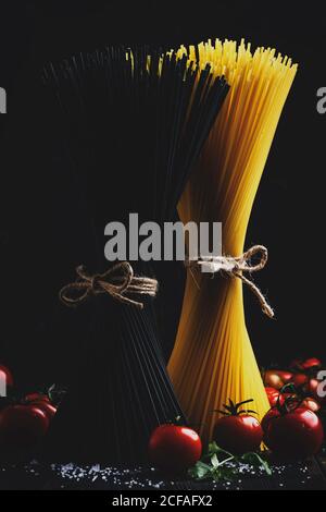 Spaghetti di pasta nero e giallo, legati con corda, si trovano verticalmente su un tavolo da cucina scuro con ingridenti per la cottura, bassa vita morta, selettivi f Foto Stock