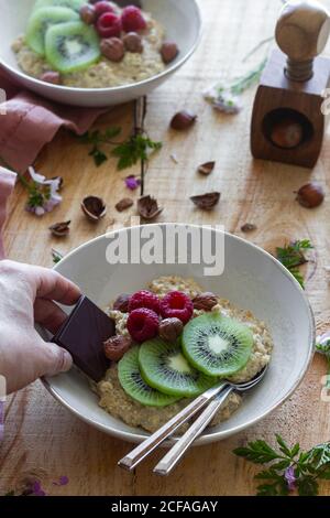 Dall'alto una persona irriconoscibile che aggiunge una fetta di cioccolato ad una ciotola sana del porridge con kiwi fresco affettato, lamponi e nocciole su un tavolo di legno con fiori Foto Stock