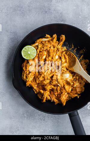 Vista dall'alto del gustoso curry tradizionale di pollo piccante con lime in padella nera con cucchiaio di legno su fondo di pietra grigia Foto Stock