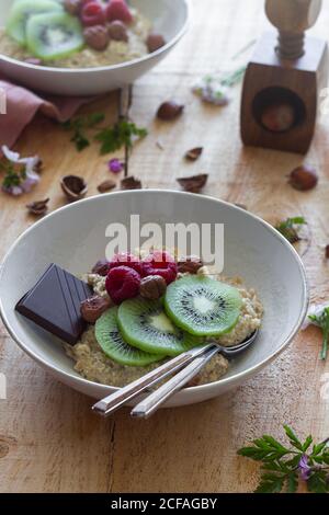 Da sopra fette di kiwi fresco e lamponi messi vicino cioccolato e nocciole in ciotola di porridge sano su legno tavolo vicino al panno lilla Foto Stock