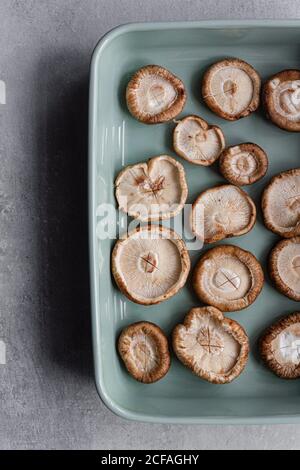 Vista dall'alto di fette di aglio aromatico su capezzoli di crudo Shiitake funghi in profondo forno di ceramica vassoio sul tavolo accanto Coltello durante la cucina gustoso piatto asiatico a casa Foto Stock