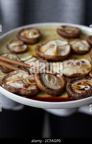 Alto angolo di funghi Shiitake cotti con aglio e soia salsa su piatto rotondo bianco con bastoncini di legno