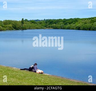 Una giovane coppia si siede sull'erba accanto all'acqua presso il lago artificiale di Ruislip Lido, Hillingdon, Londra nord-occidentale Foto Stock