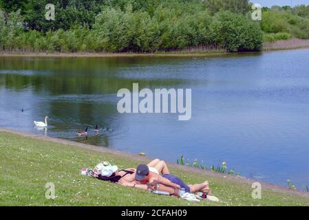 La giovane coppia si trova sull'erba accanto all'acqua al lago artificiale di Ruislip Lido con scarpe e giacche staccate. Swan & oche nuotare in lago. Londra nord-occidentale. Foto Stock
