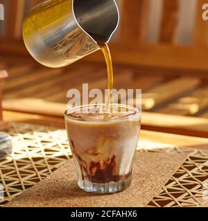 persona che versa una gustosa bevanda calda dalla caffettiera metallica in bicchiere trasparente con latte fresco e ghiaccio durante la preparazione della colazione a casa Foto Stock