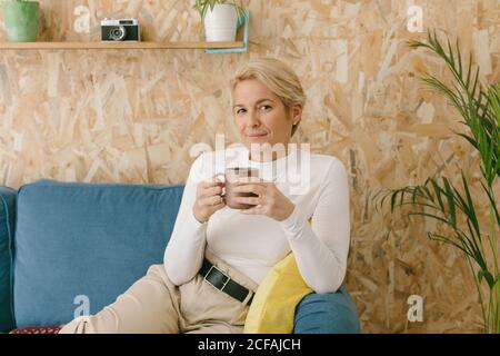 Donna d'affari calma adulto con i capelli corti biondi seduti sul divano accogliente in ufficio avendo tazza di caffè e sorridendo serenamente alla macchina fotografica Foto Stock