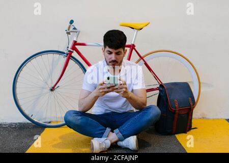 uomo che controlla il suo smartphone. È seduto sul pavimento con una bicicletta e uno zaino. Foto Stock