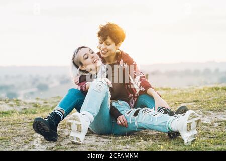 Ridendo le amiche strette in abiti casual eleganti seduti sul verde erba in abbraccio con cielo su sfondo Foto Stock