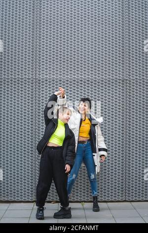 Tutta la lunghezza dei soddisfare allegri amici multietnici femminili in stile abiti caldi che ballano in piedi e sorridono con un edificio moderno sfondo Foto Stock