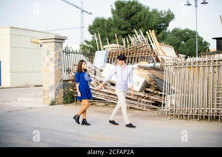 Uomo elegante e donna affascinante in vestito blu tenendo le mani e camminare per strada lungo le case in ricostruzione Foto Stock