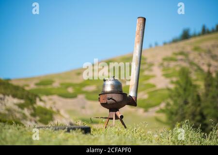 Stufa a legna arrugginita con bollitore di metallo posto sullo sfondo di paesaggio pittoresco con terreno erboso e cielo blu Foto Stock