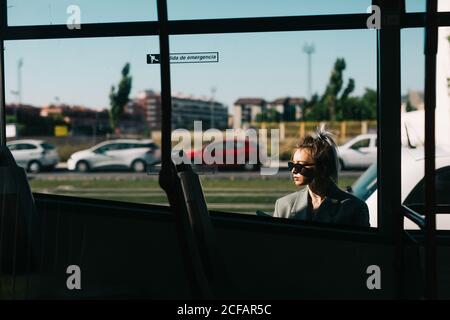 Donna d'affari alla moda e occhiali da sole appoggiati dietro l'autobus e guardando lontano Foto Stock