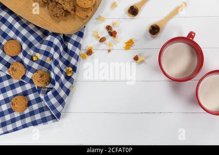 Vista dei biscotti, cucchiai con frutta secca e noci e due tazze con latte su legno Foto Stock