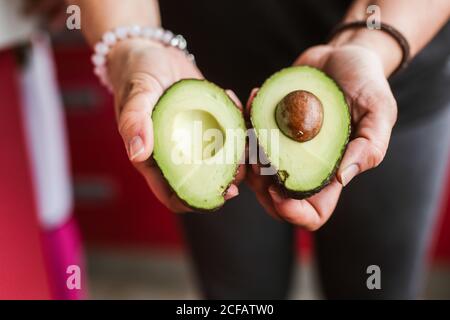 donna irriconoscibile che dimostra due metà di avocado maturo alla macchina fotografica mentre si è in piedi su sfondo sfocato in cucina Foto Stock