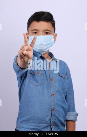 Ragazzo indiano carino e intelligente in maschera chirurgica che mostra la vittoria Pose.ITS una foto frontale in cui il ragazzo sembra sicuro e. esprimersi con la mano Foto Stock