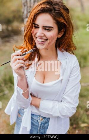 Donna attraente con pennello sorridente e guardando via mentre spende il tempo e dipingere in natura Foto Stock