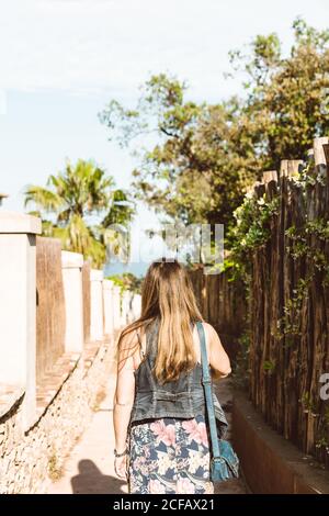 Vista sul retro di una donna irriconoscibile che cammina sulla strada stretta sotto il sole Foto Stock