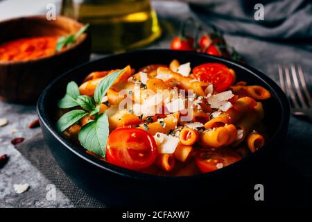 Deliziosa pasta decorata con pomodori ciliegini maturi e basilico fresco Vicino alla ciotola di legno con salsa arrabiata e parmigiano grattugiato Foto Stock