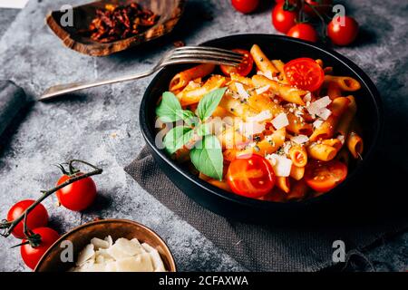 Da sopra deliziosa pasta decorata con pomodori ciliegini maturi e. basilico fresco vicino a ciotola di legno con salsa di arrabiata e grattugiato Parmigiano Foto Stock