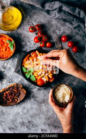 Dall'alto di cuoco anonimo aggiungere il parmigiano grattugiato a. gustosa pasta fatta in casa con pomodori freschi e basilico sul tavolo in cucina Foto Stock