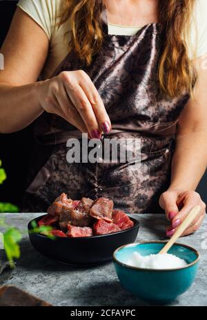 Donna senza volto che indossa grembiule in piedi a tavola e aggiungendo sale in ciotola con fette di manzo crudo mentre prepara la carne per la cottura Foto Stock