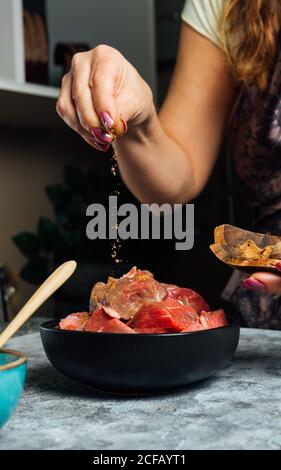 Donna senza volto che indossa grembiule in piedi a tavola e aggiungendo sale in ciotola con fette di manzo crudo mentre prepara la carne per la cottura Foto Stock