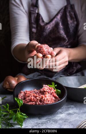 Da sopra persona irriconoscibile che indossa un grembiule mostrando polpette alla fotocamera sopra le ciotole con carne macinata durante la preparazione del pranzo Foto Stock