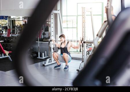 Potente sportivo determinato che fa esercizio con il barbell a squat rack durante l'allenamento di sollevamento pesi con allenatore personale in palestra moderna Foto Stock