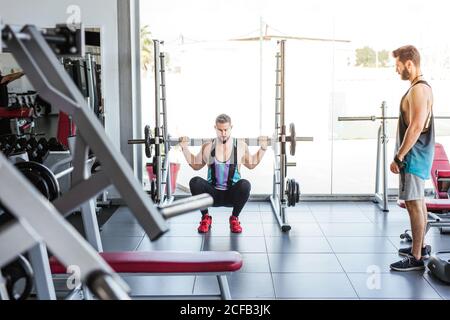 Potente sportivo determinato che fa esercizio con il barbell a squat rack durante l'allenamento di sollevamento pesi con allenatore personale in palestra moderna Foto Stock