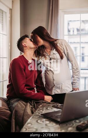 Giovane uomo in ponticello rosso seduto sul divano e baciando moglie incinta che si trova nelle vicinanze mentre si passa del tempo libero a casa e godendo del tempo insieme Foto Stock