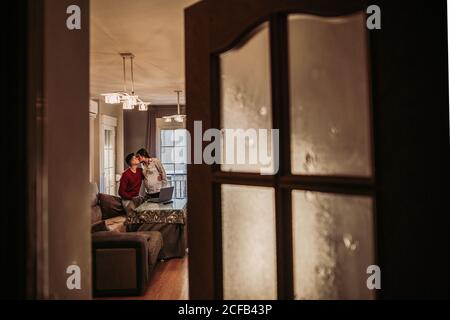 Giovane uomo in ponticello rosso seduto sul divano e baciando moglie incinta che si trova nelle vicinanze mentre si passa del tempo libero a casa e godendo del tempo insieme Foto Stock