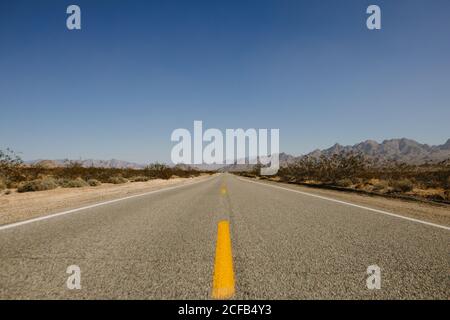 Strada asfaltata rettilinea che attraversa terreni sabbiosi con erba secca e le montagne sullo sfondo del cielo nuvoloso in giorno di caldo In California Foto Stock