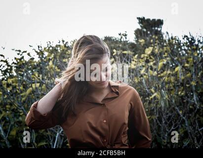 Allegra bella donna in piedi di fronte a cespugli che toccano il retro della testa con la mano che guarda verso il basso Foto Stock