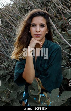 Elegante ragazza capelli ricci turista in camicia verde scuro e. body muta guardando la macchina fotografica mentre camminando sulla foresta con fico albero sullo sfondo Foto Stock