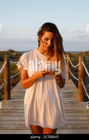 Pensivo capelli lunghi Donna surf cellulare mentre cammina su ponte sospeso in luce del sole Foto Stock
