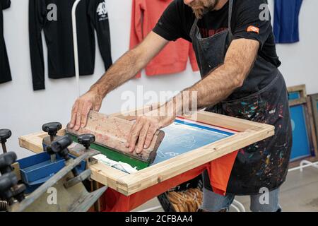 Crop uomo in grembiule sporco lavorando con serigrafia durante la creazione stampa su t-shirt in officina Foto Stock