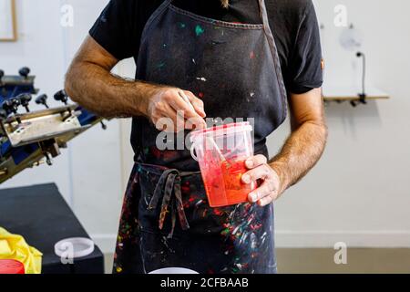 Raccolto concentrato uomo che indossa grembiule sporco che mescola varie vernici per serigrafia in officina Foto Stock