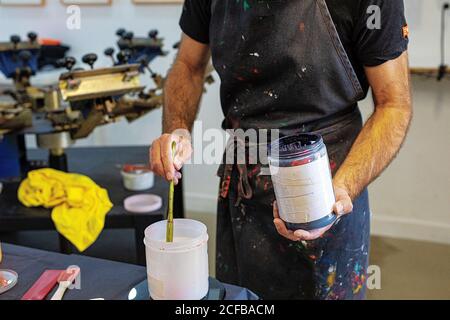 Raccolto concentrato uomo che indossa grembiule sporco che mescola varie vernici per serigrafia in officina Foto Stock